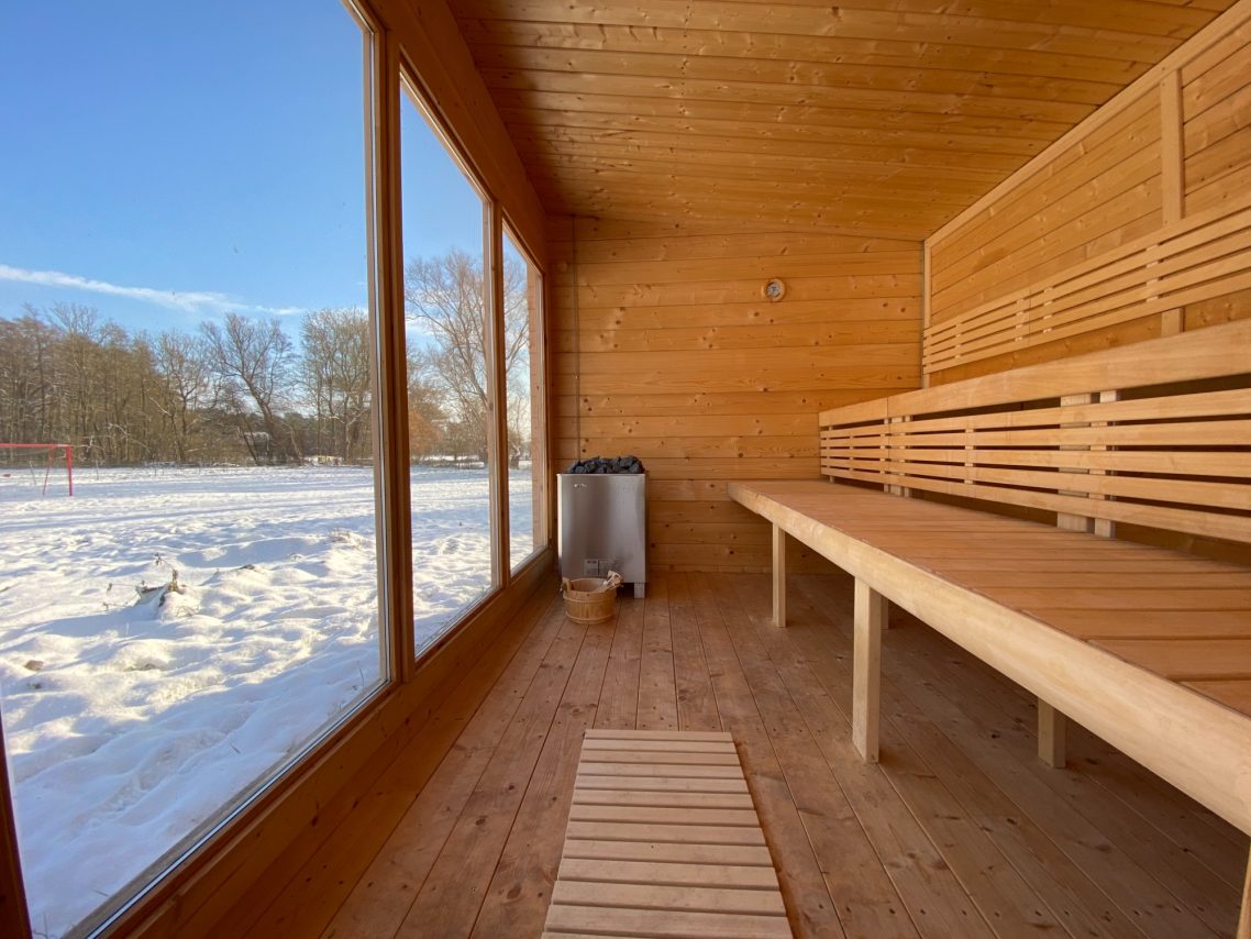 Mach's dir in unserer Sauna gemütlich. Innenansicht einer Sauna mit Holzbänken und großen Fenstern, im Hintergrund Schnee.