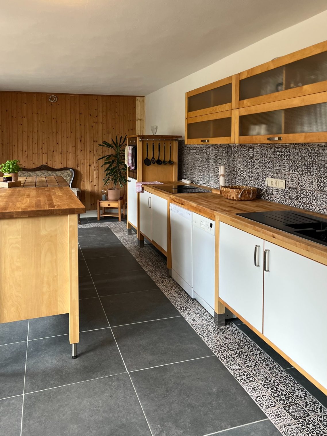 Zwei Geschirrspüler, ein großes Induktionskochfeld und ein separater Backofen Moderne Küche mit Holzoberflächen, grauen Fliesen und Pflanzen im Hintergrund.