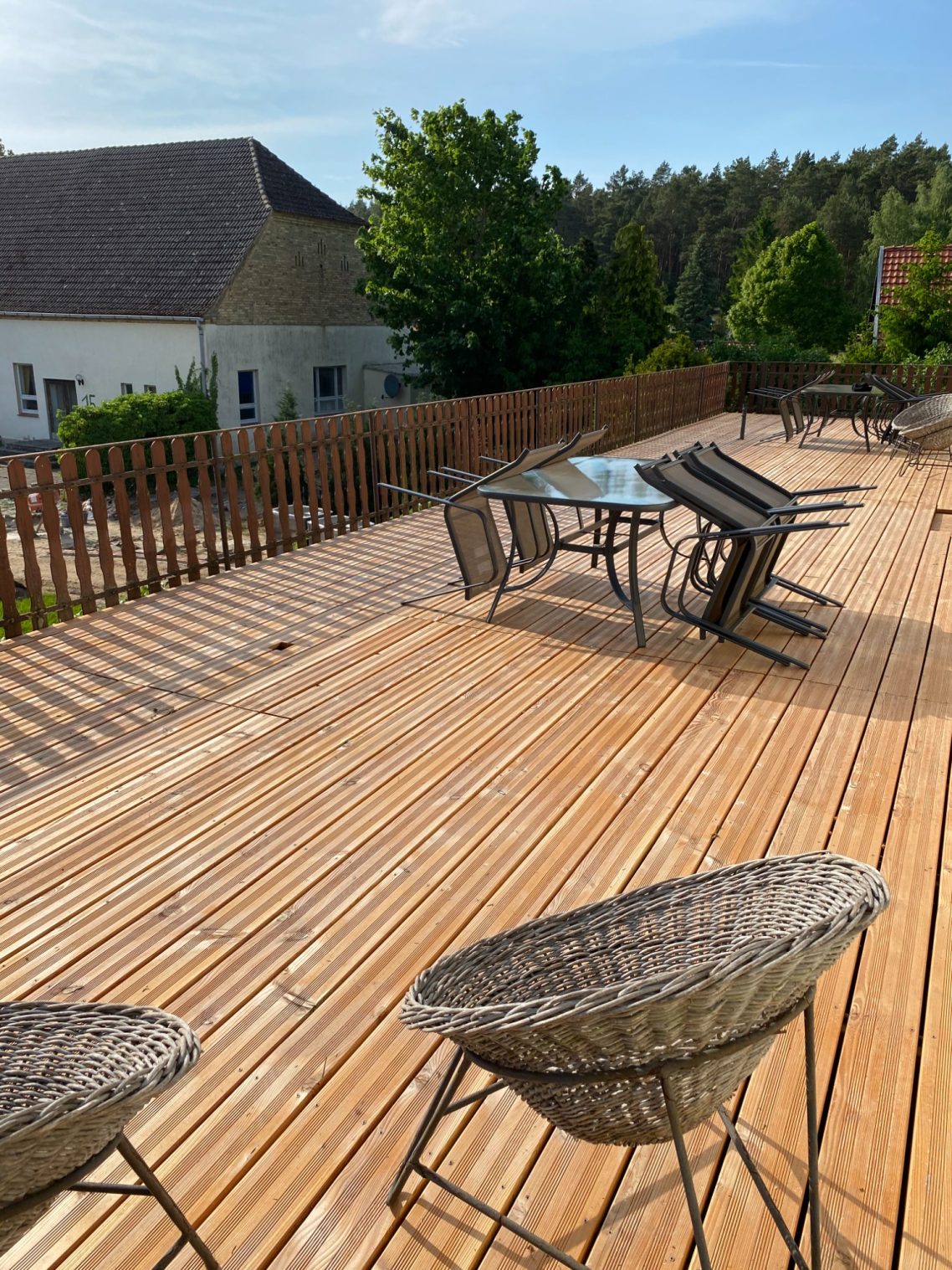 Sonnenterrasse Holzterrasse mit Stühlen und einem Tisch, im Hintergrund ein Haus und Bäume.