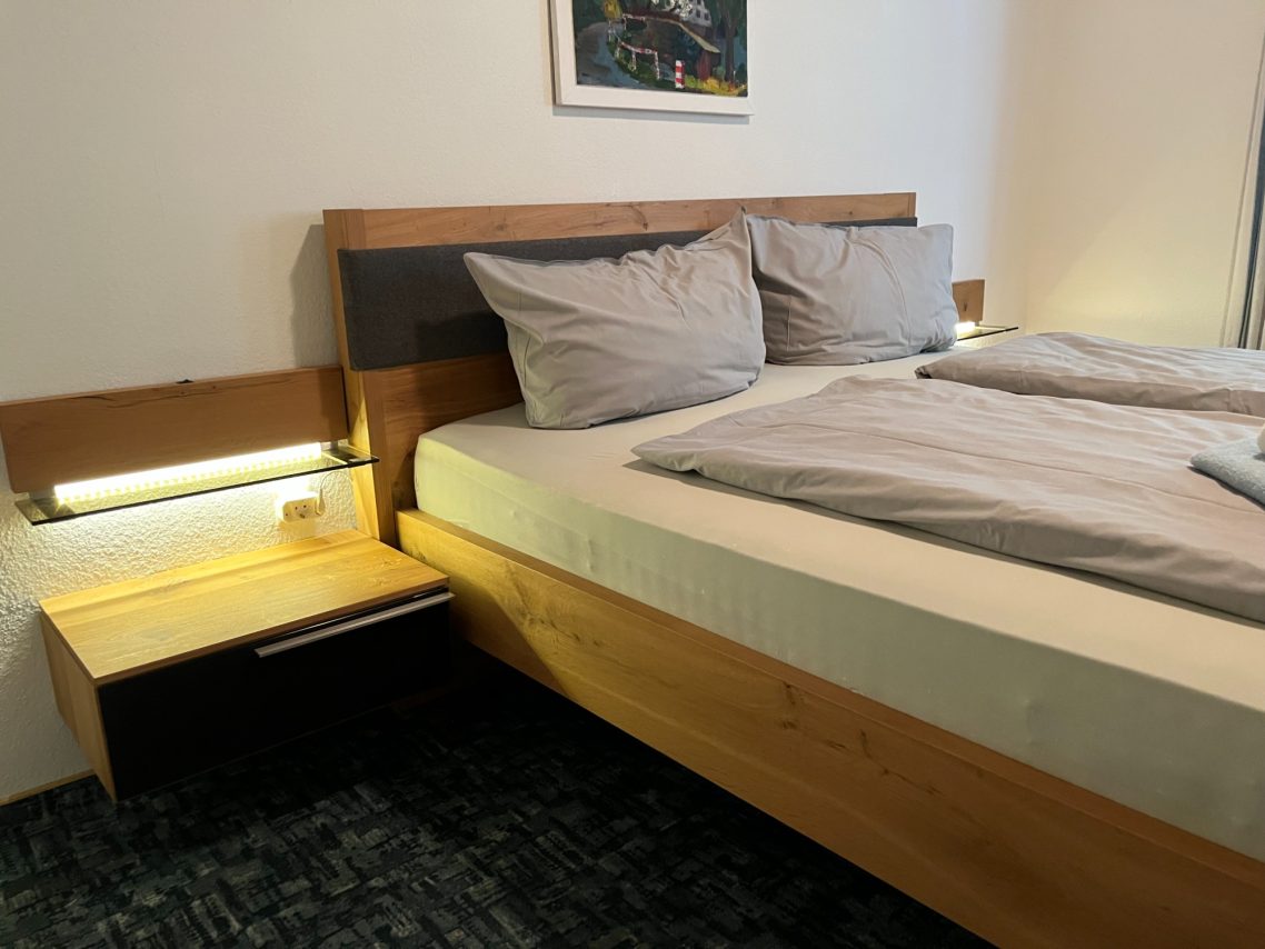 Bett mit zwei Kissen, Nachttisch und modernen Leuchten in einem hellen Schlafzimmer.
